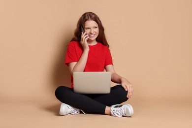 Smiling teenage girl with laptop talking by smartphone on beige background