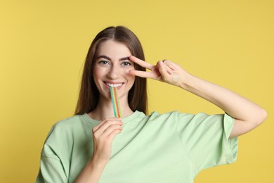 Photo of Young woman eating tasty rainbow sour belt while showing v-sign on yellow background