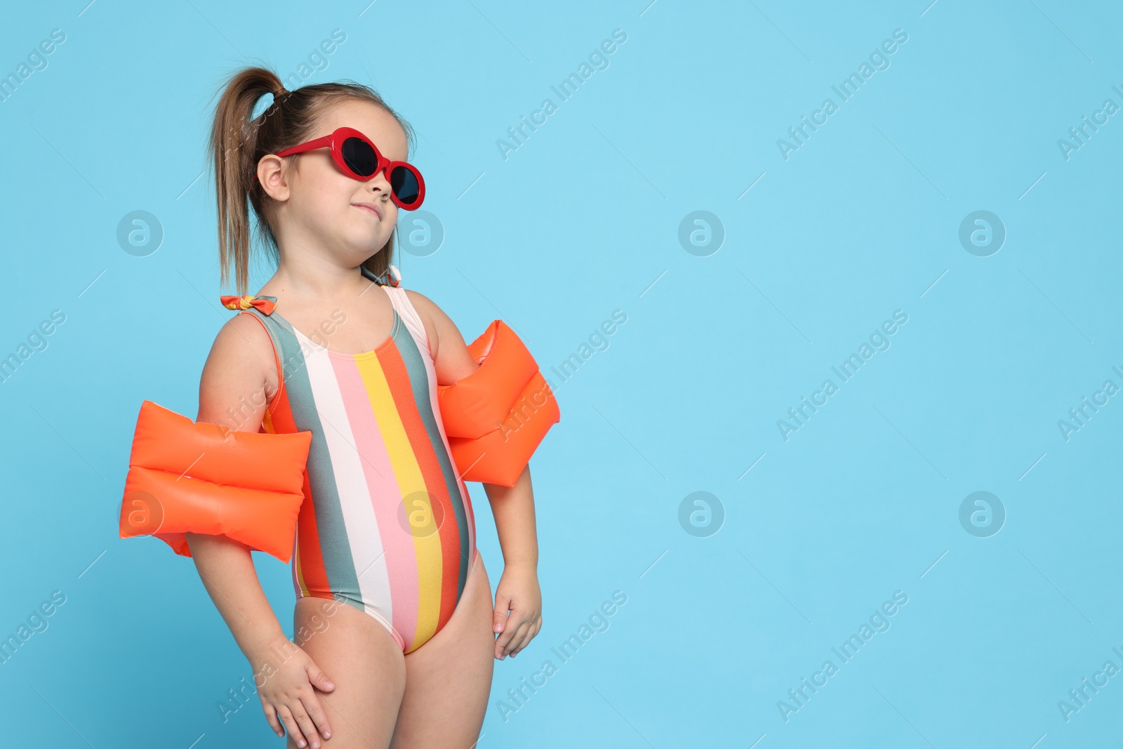 Photo of Cute little girl in swimsuit with swim armbands on light blue background. Space for text