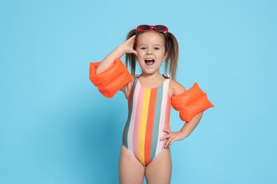 Photo of Cute little girl in swimsuit with swim armbands on light blue background