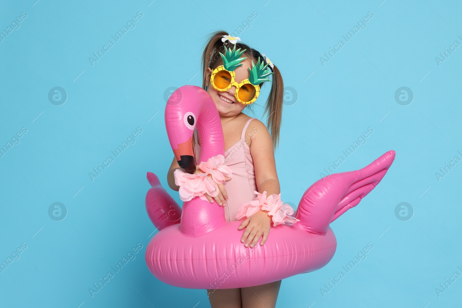 Photo of Cute little girl in swimsuit with funny sunglasses and inflatable ring on light blue background