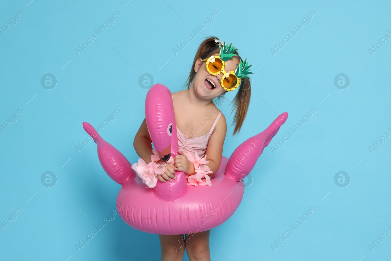 Photo of Cute little girl in swimsuit with funny sunglasses and inflatable ring on light blue background