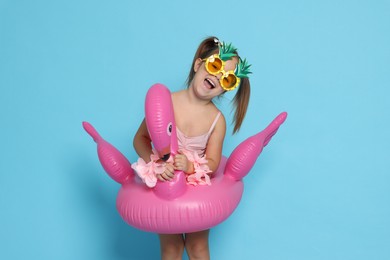 Photo of Cute little girl in swimsuit with funny sunglasses and inflatable ring on light blue background