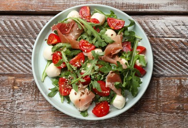 Photo of Tasty salad with arugula, mozzarella cheese, tomatoes and jamon on wooden table, top view