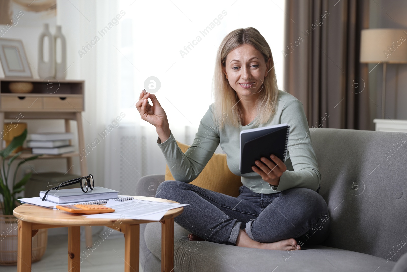 Photo of Budget planning. Woman with notebook and pen on sofa indoors