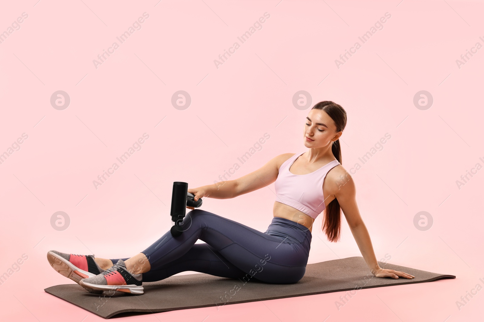 Photo of Woman using percussive massager to relax leg muscles on mat against pink background