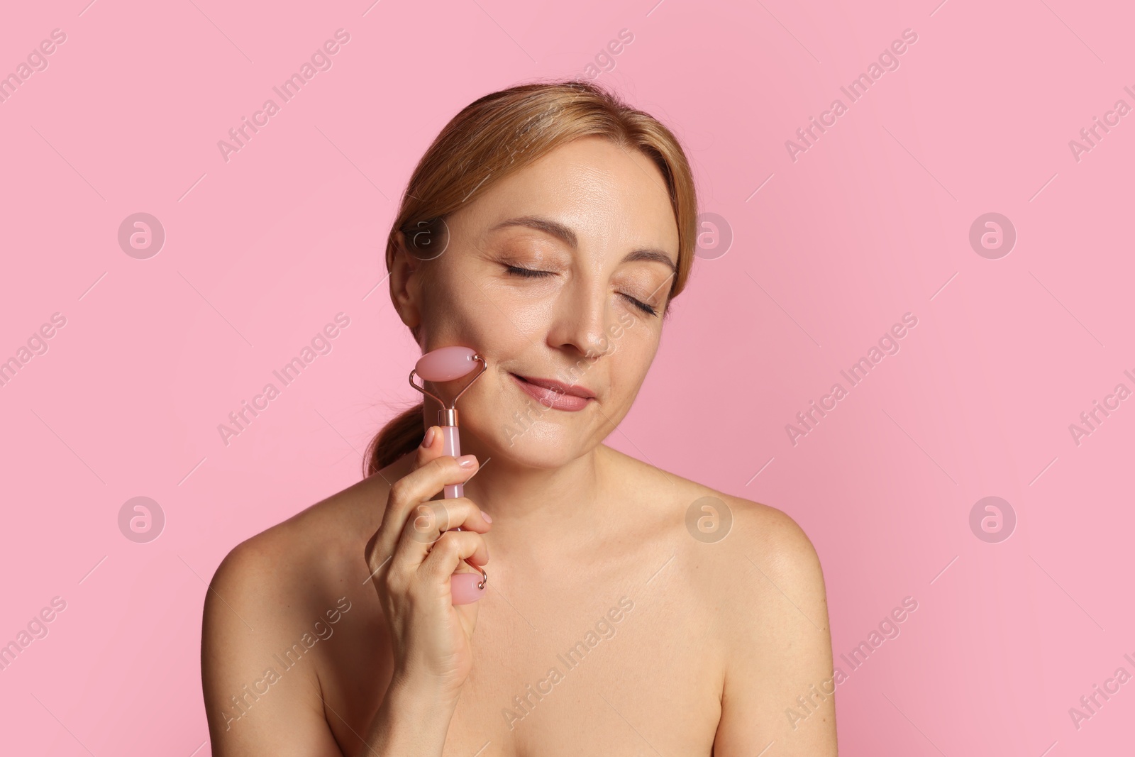 Photo of Beautiful woman doing facial self massage with roller on pink background