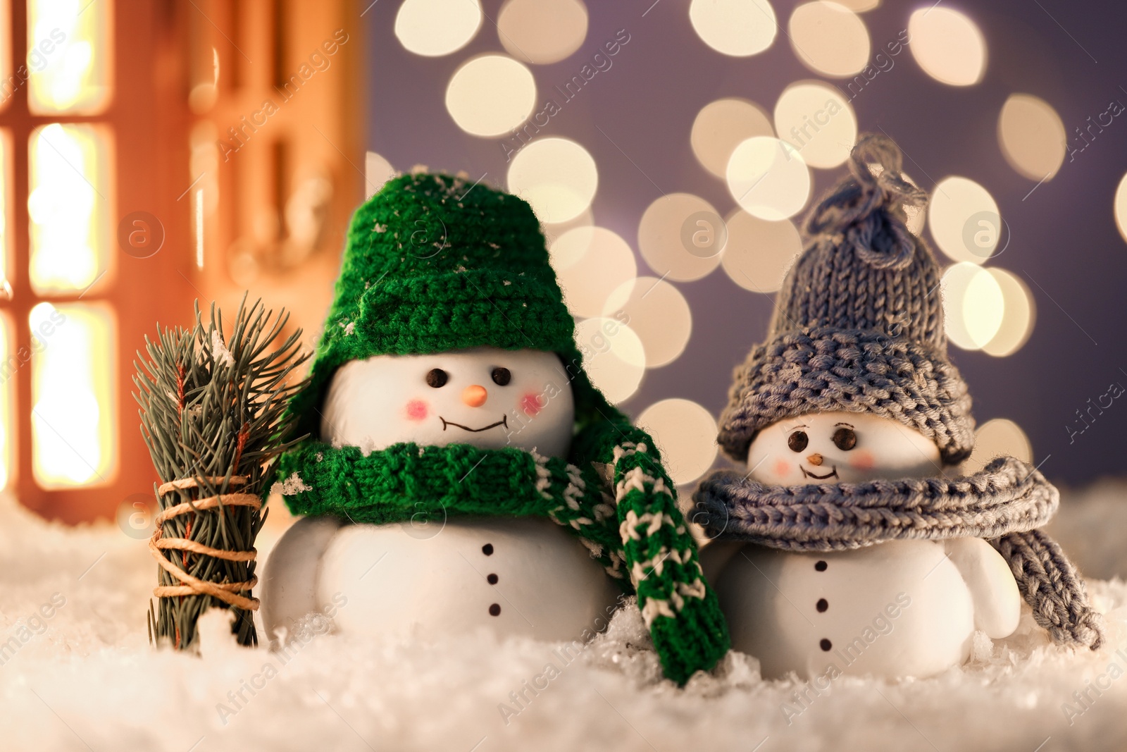 Photo of Cute decorative snowmen and fir tree branches on artificial snow against blurred lights, closeup