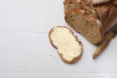 Photo of Fresh bread with butter and knife on white tiled table, flat lay. Space for text