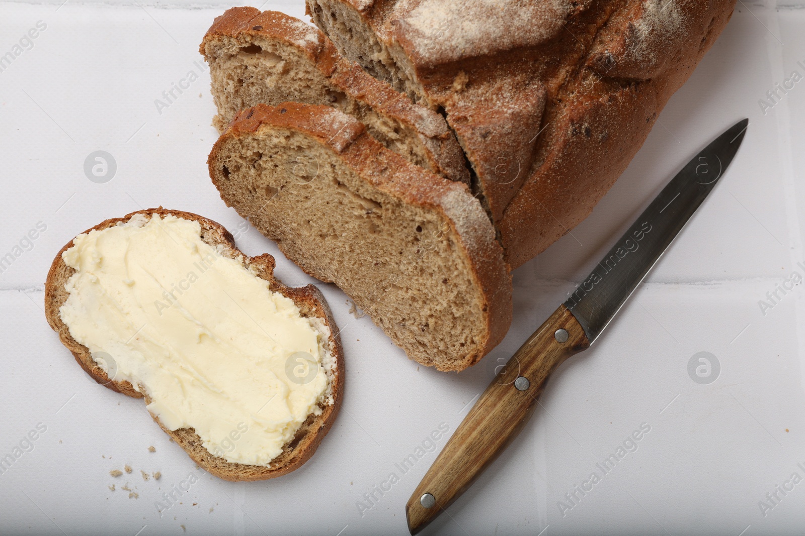 Photo of Fresh bread with butter and knife on white tiled table, top view