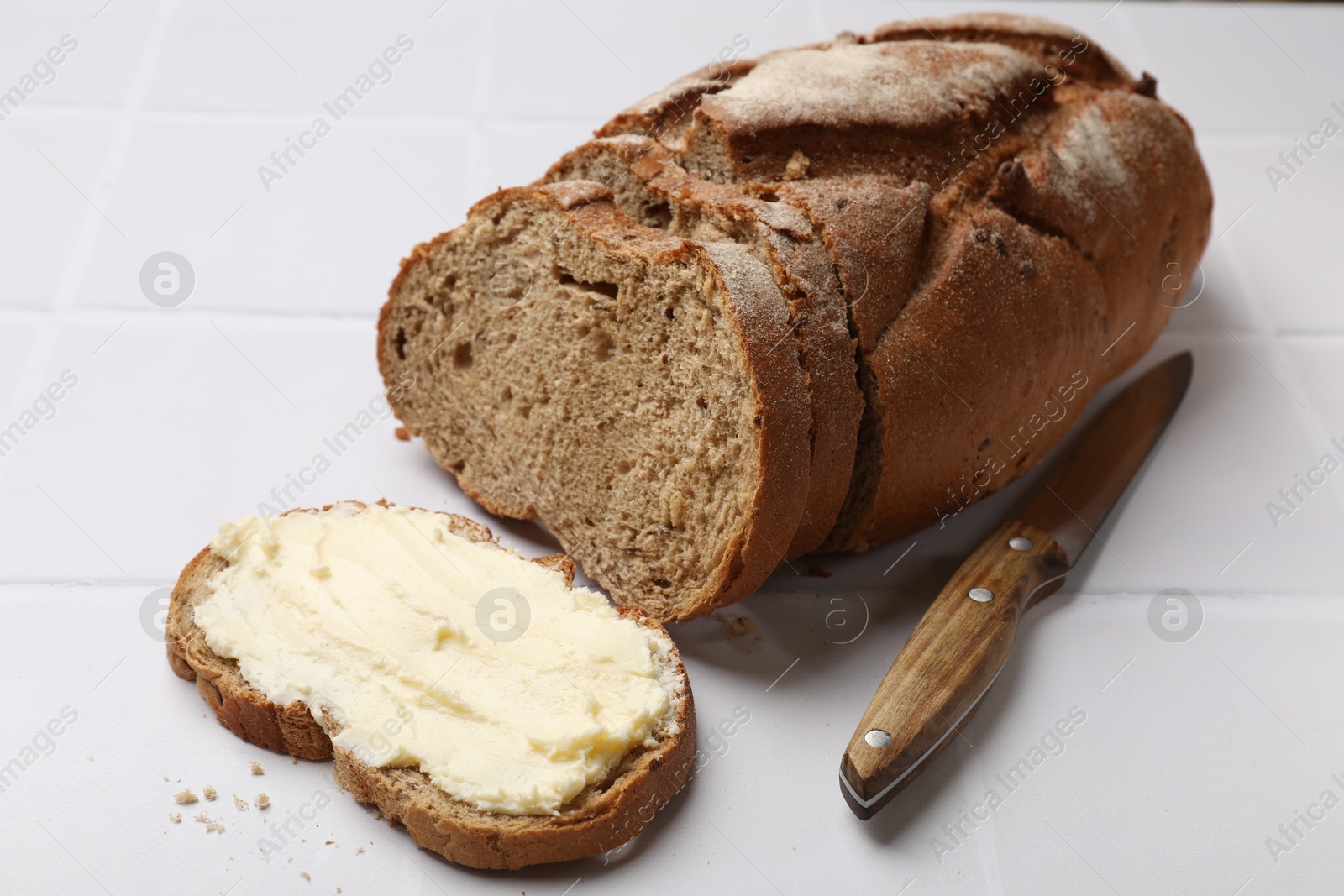 Photo of Fresh bread with butter and knife on white tiled table, closeup