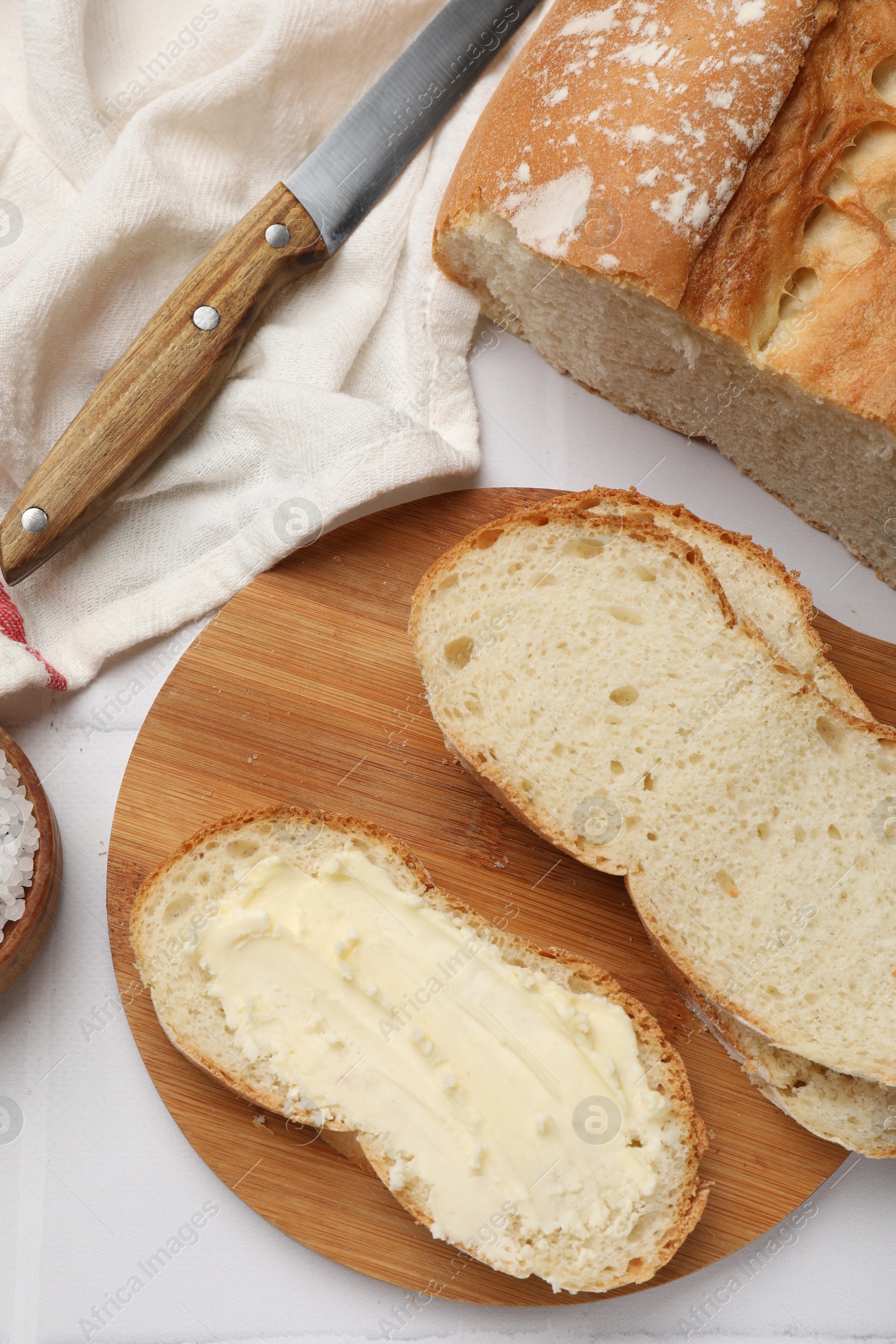 Photo of Fresh bread with butter and knife on white tiled table, flat lay