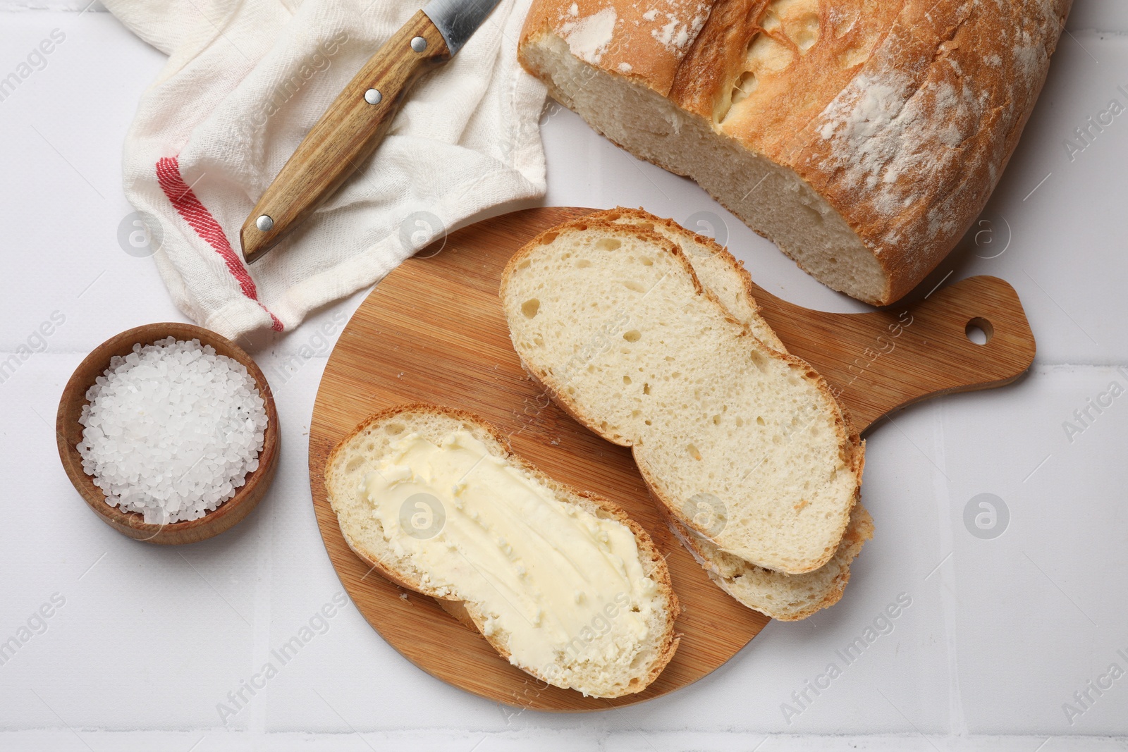 Photo of Fresh bread with butter and knife on white tiled table, flat lay