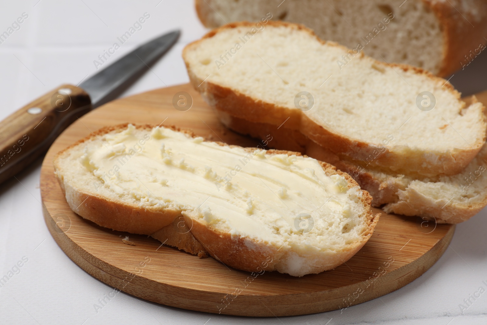 Photo of Fresh bread with butter and knife on white tiled table, closeup