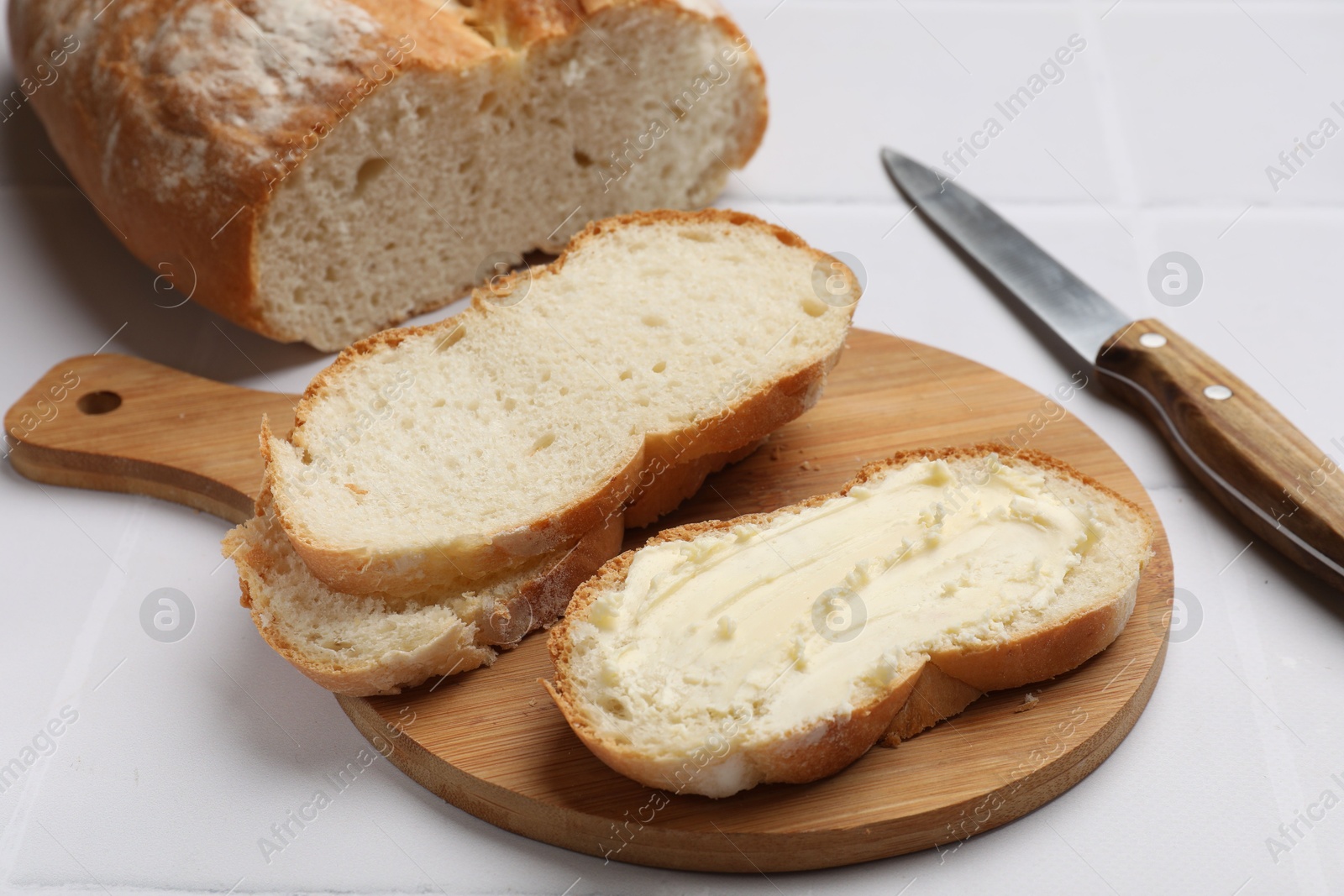 Photo of Fresh bread with butter and knife on white tiled table, closeup