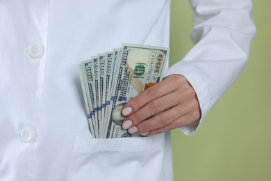 Photo of Corruption concept. Doctor putting dollar banknotes into her pocket on green background, closeup