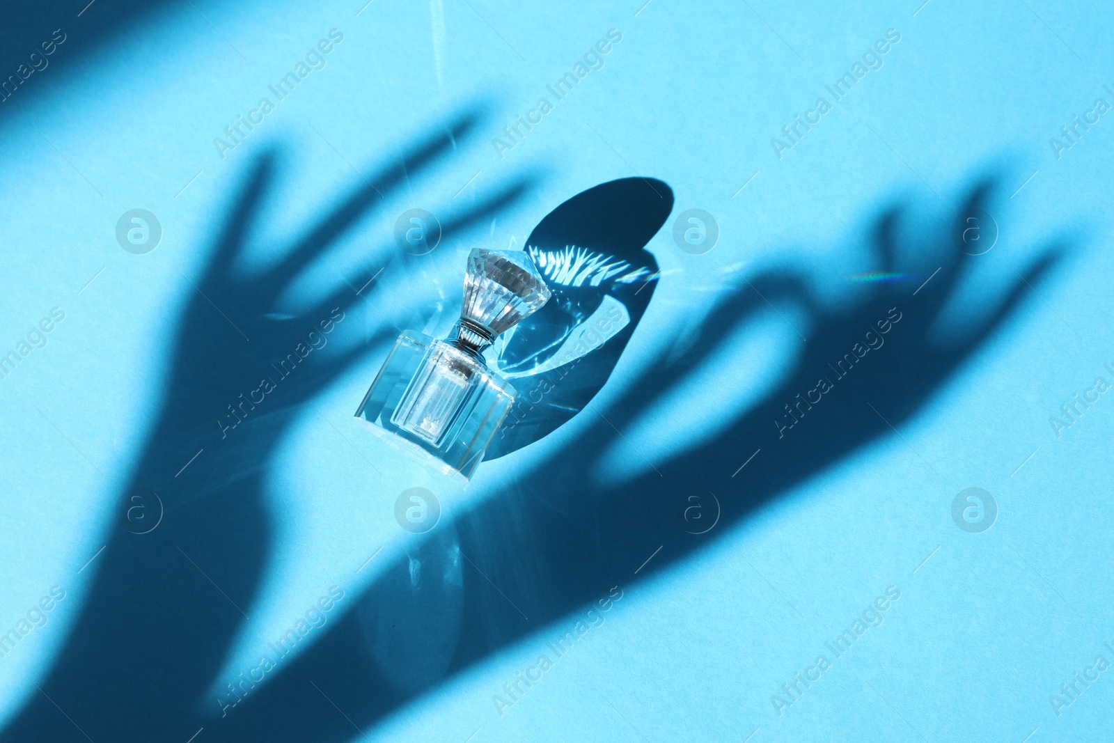 Photo of Perfume bottle and shadow of woman's hands on light blue background, top view