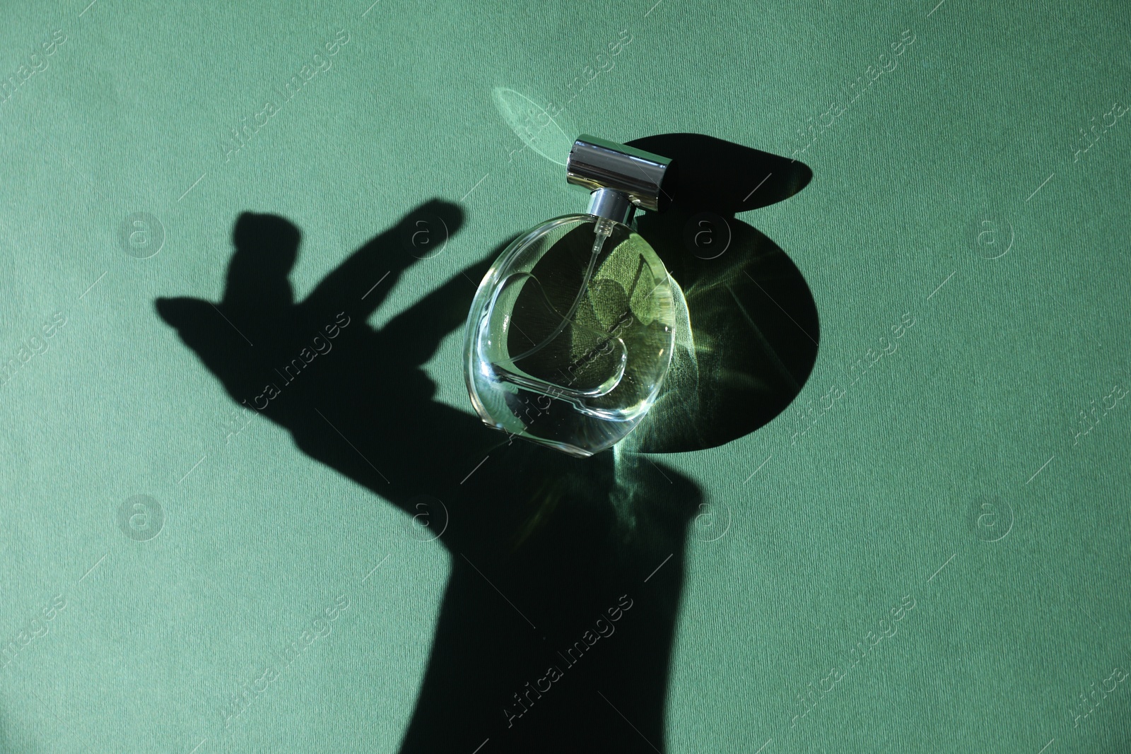 Photo of Perfume bottle and shadow of woman's hand on green background, top view