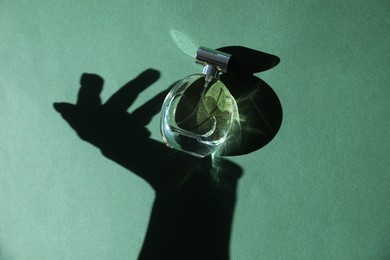 Perfume bottle and shadow of woman's hand on green background, top view