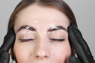 Photo of Brow lamination. Cosmetologist putting film onto woman's eyebrows against grey background, closeup