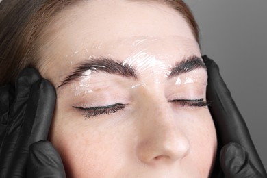 Photo of Brow lamination. Cosmetologist putting film onto woman's eyebrows against grey background, closeup