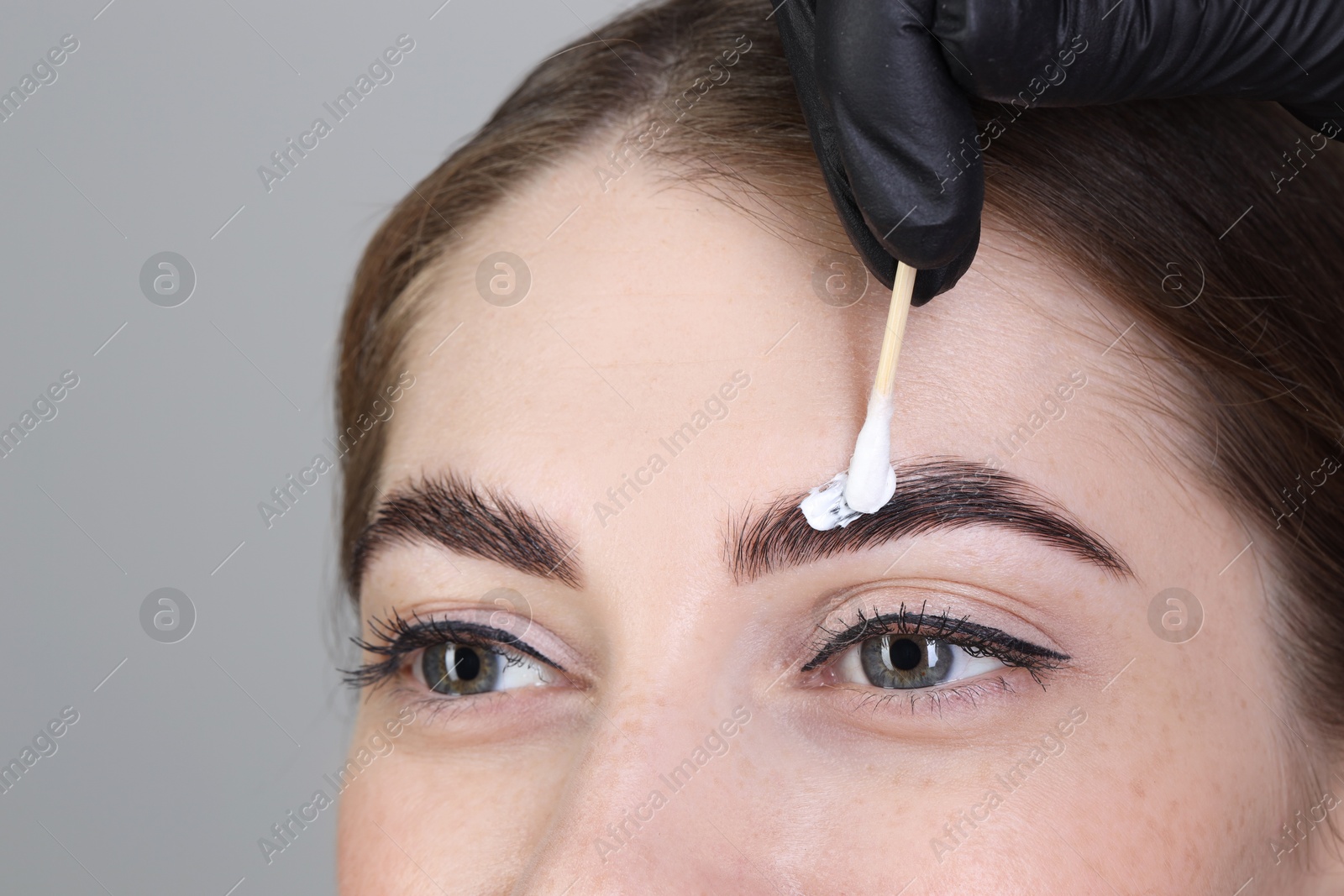 Photo of Brow lamination. Cosmetologist applying cream onto woman's eyebrows against grey background, closeup