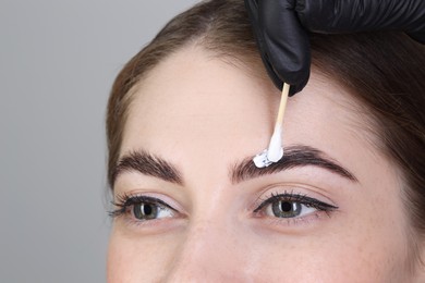 Photo of Brow lamination. Cosmetologist applying cream onto woman's eyebrows against grey background, closeup