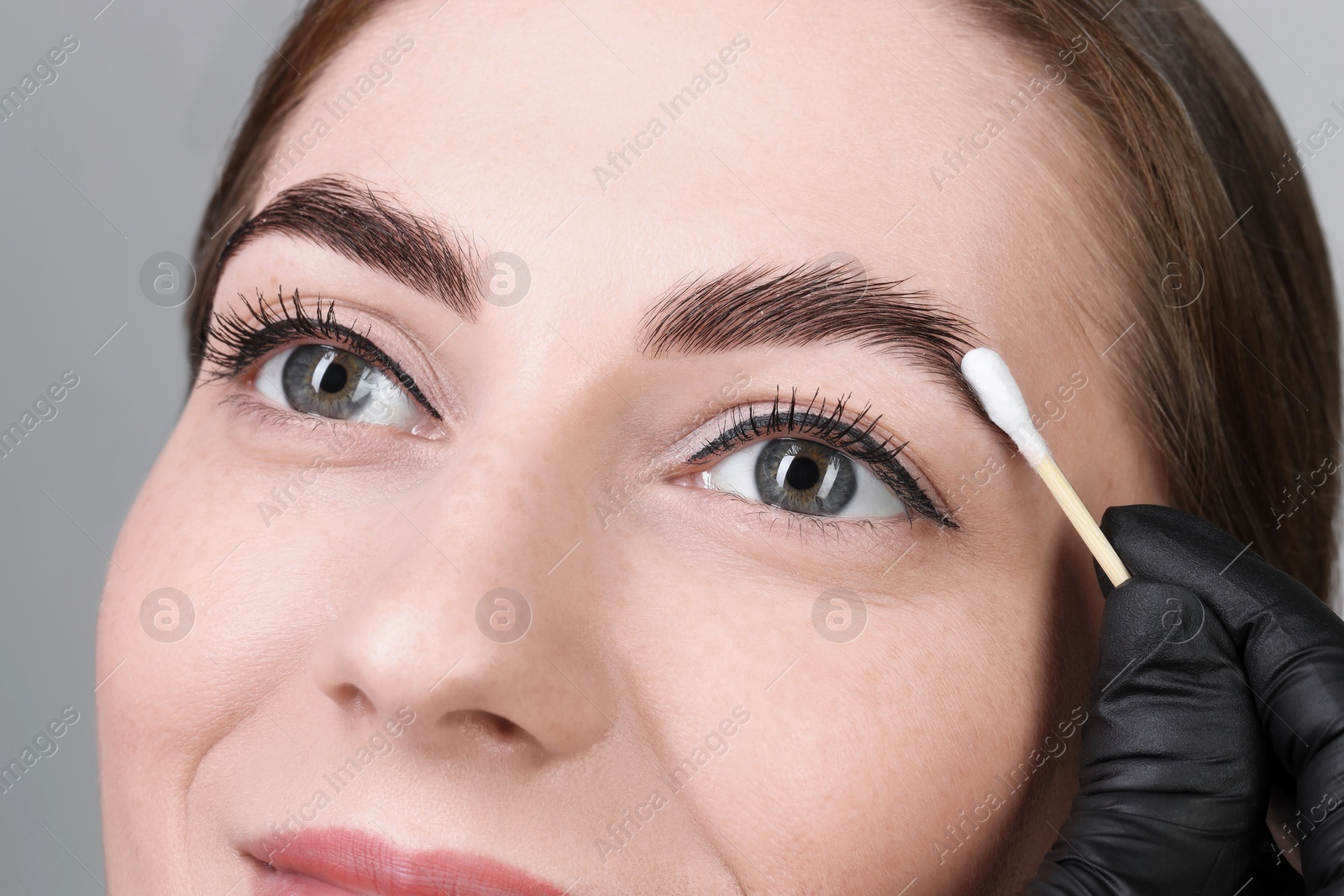 Photo of Brow lamination. Cosmetologist applying cream onto woman's eyebrows against grey background, closeup