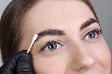 Photo of Brow lamination. Cosmetologist applying cream onto woman's eyebrows against grey background, closeup