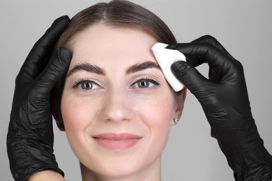 Photo of Brow lamination. Cosmetologist wiping woman's eyebrows with cotton pad against grey background, closeup