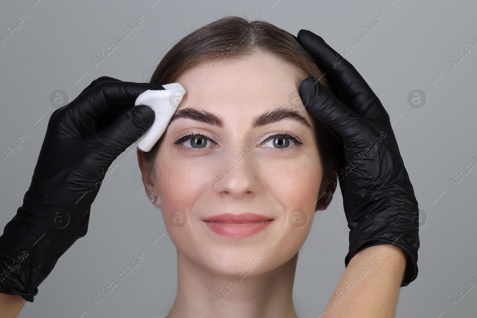 Photo of Brow lamination. Cosmetologist wiping woman's eyebrows with cotton pad against grey background, closeup