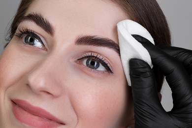 Brow lamination. Cosmetologist wiping woman's eyebrows with cotton pad against grey background, closeup