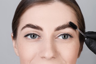 Photo of Brow lamination. Cosmetologist combing woman's eyebrows with brush against grey background, closeup