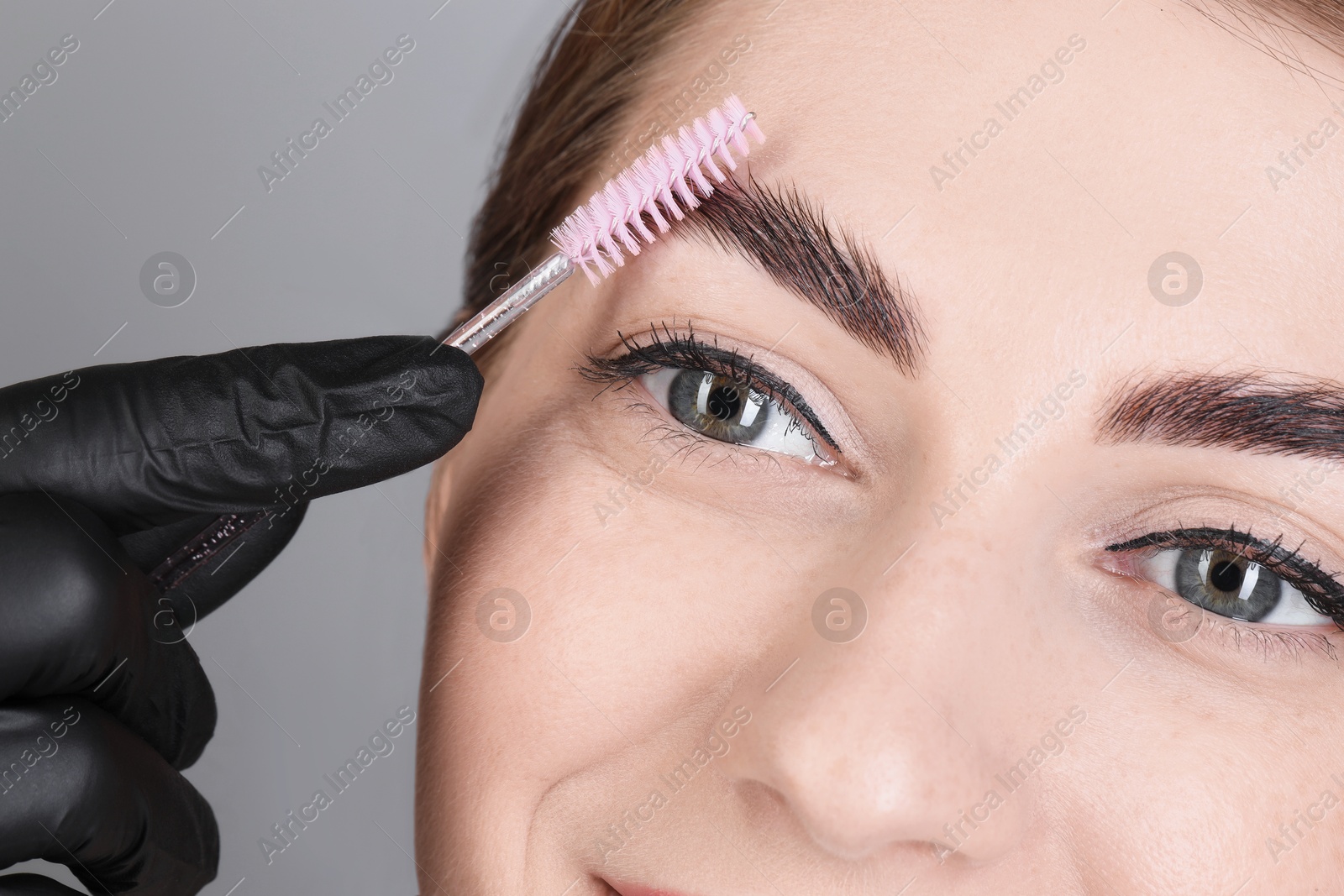 Photo of Brow lamination. Cosmetologist combing woman's eyebrows with brush against grey background, closeup