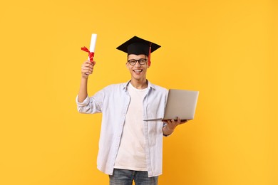 Happy student with diploma and laptop after graduation on orange background