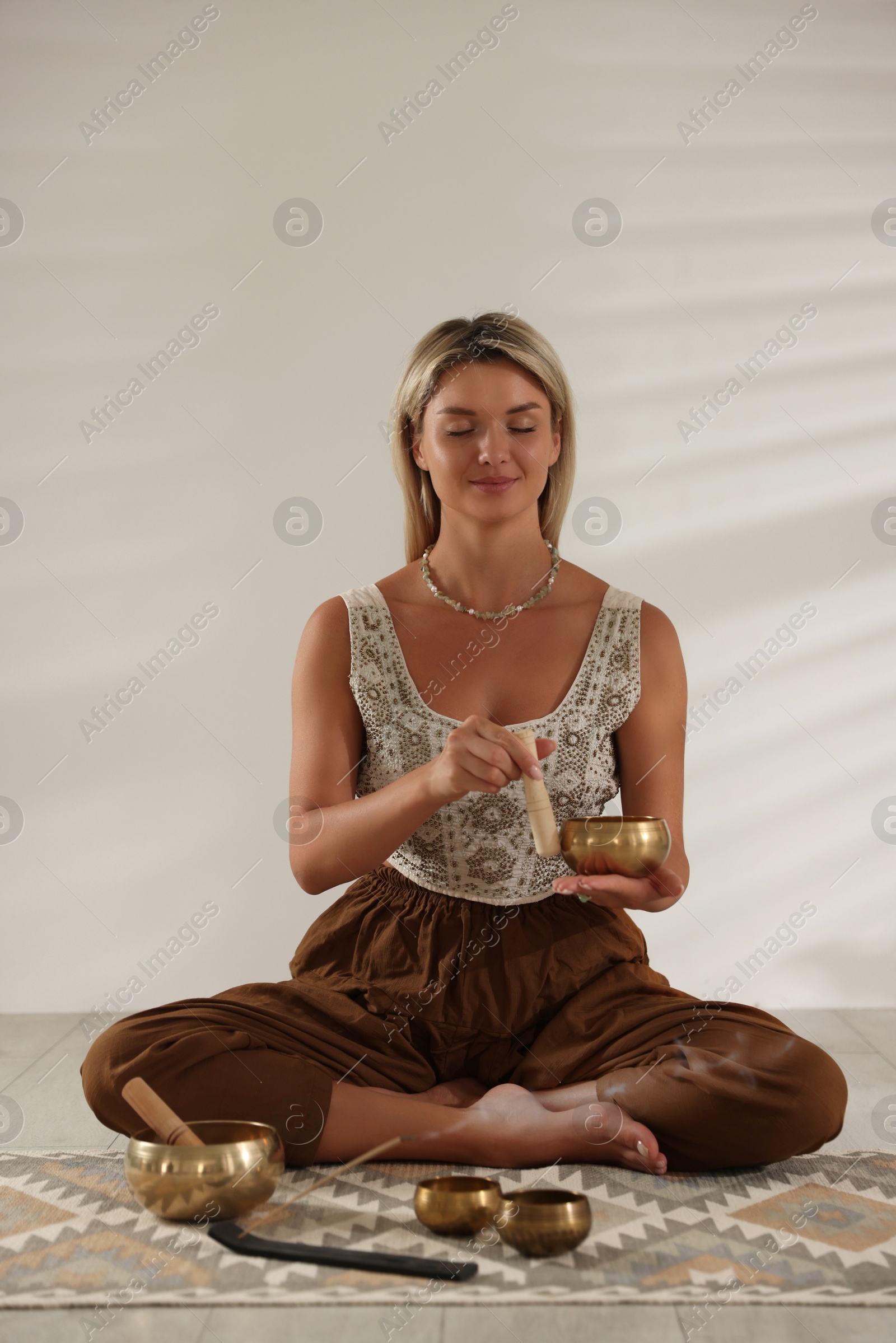 Photo of Beautiful young woman with smoldering incense stick and tibetan singing bowls on floor indoors