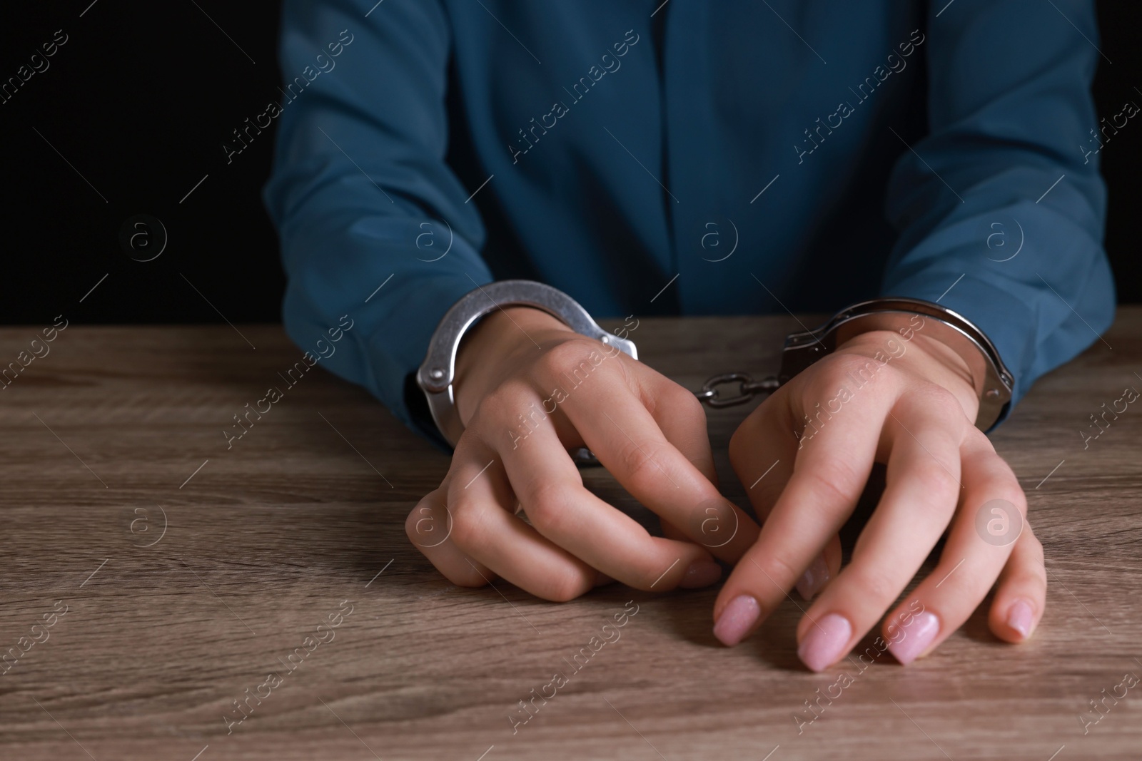 Photo of Woman in metal handcuffs at wooden table against black background, closeup