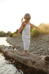 Photo of Cute little girl walking along tree trunk near river. Child enjoying beautiful nature