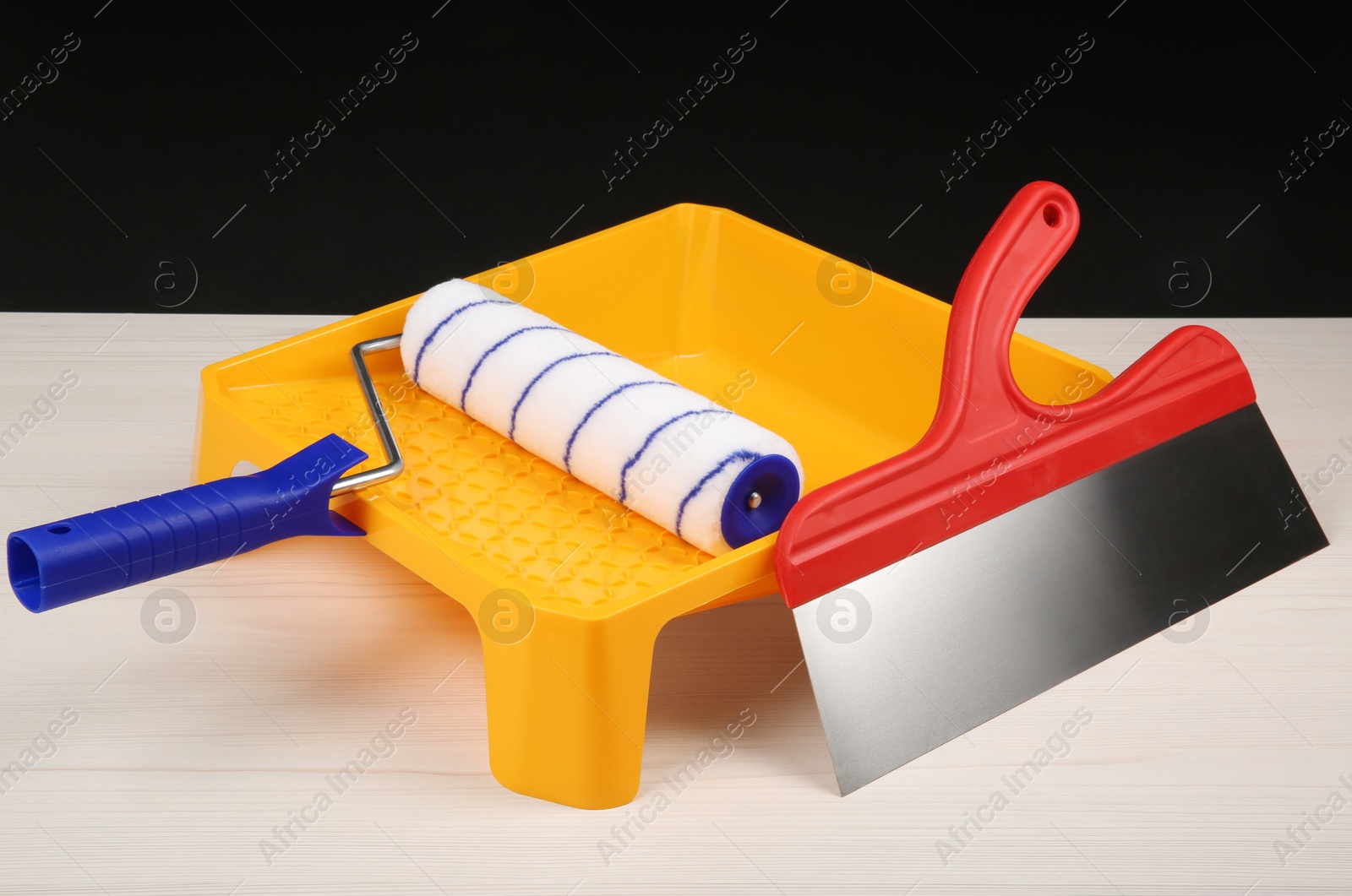 Photo of Paint roller, tray and putty knife on light wooden table against black background. Construction tools