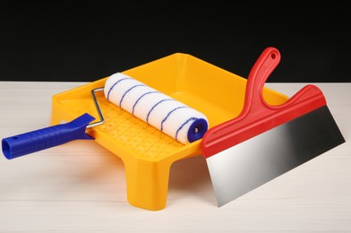 Photo of Paint roller, tray and putty knife on light wooden table against black background. Construction tools