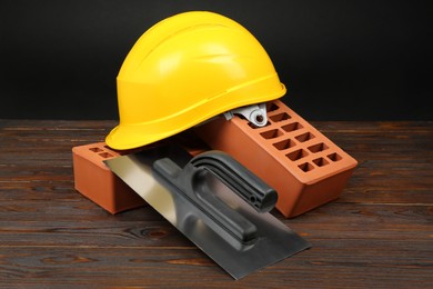 Photo of Red bricks, putty knife and yellow hard hat on wooden table. Building material