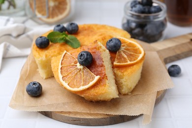 Photo of Pieces of delicious semolina cake with blueberries and orange slices on white tiled table, closeup