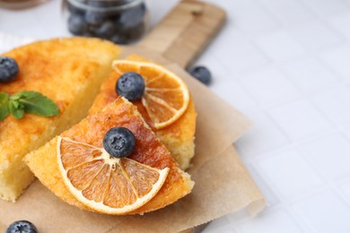 Photo of Pieces of delicious semolina cake with blueberries and orange slices on white tiled table, closeup. Space for text