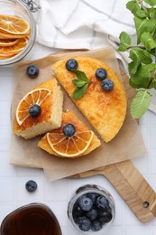 Photo of Pieces of delicious semolina cake with blueberries and orange slices on white tiled table, flat lay