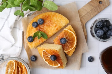 Photo of Pieces of delicious semolina cake with blueberries and orange slices on white tiled table, flat lay