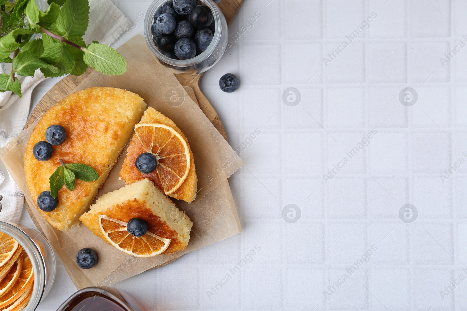 Photo of Pieces of delicious semolina cake with blueberries and orange slices on white tiled table, flat lay. Space for text