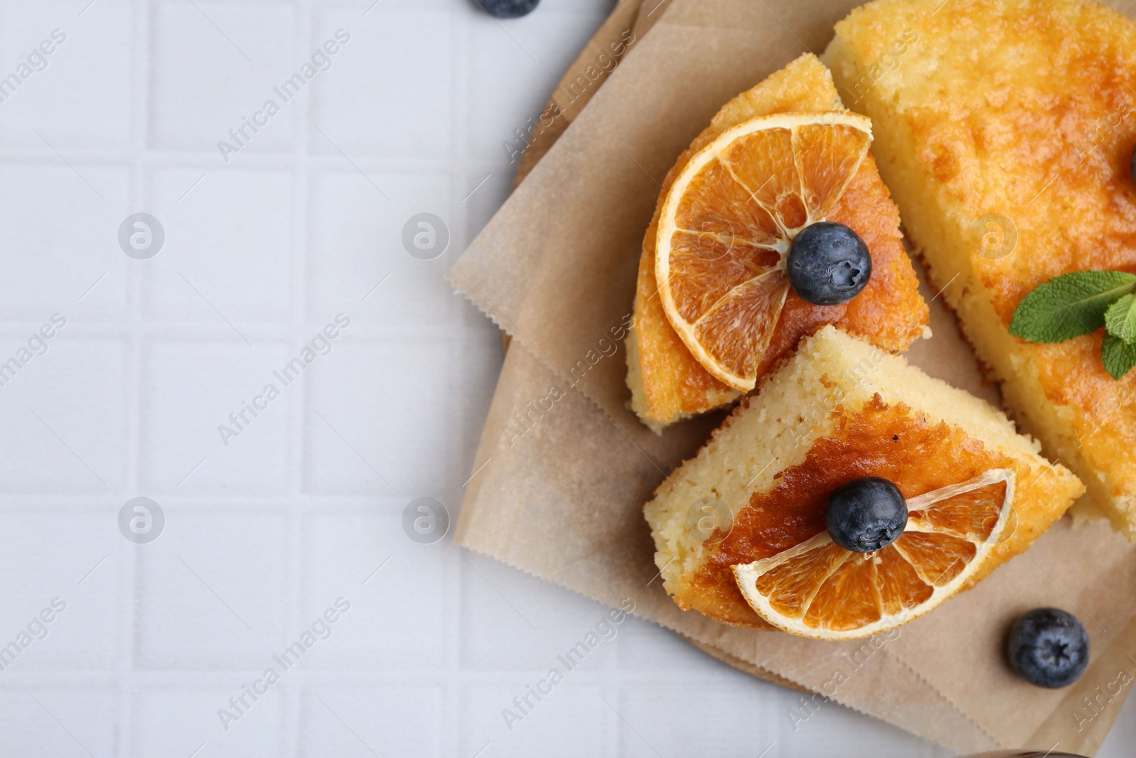 Photo of Pieces of delicious semolina cake with blueberries and orange slices on white tiled table, top view. Space for text