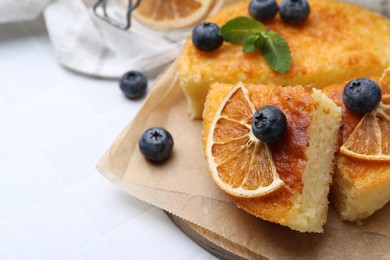 Photo of Pieces of delicious semolina cake with blueberries and orange slices on white tiled table, closeup