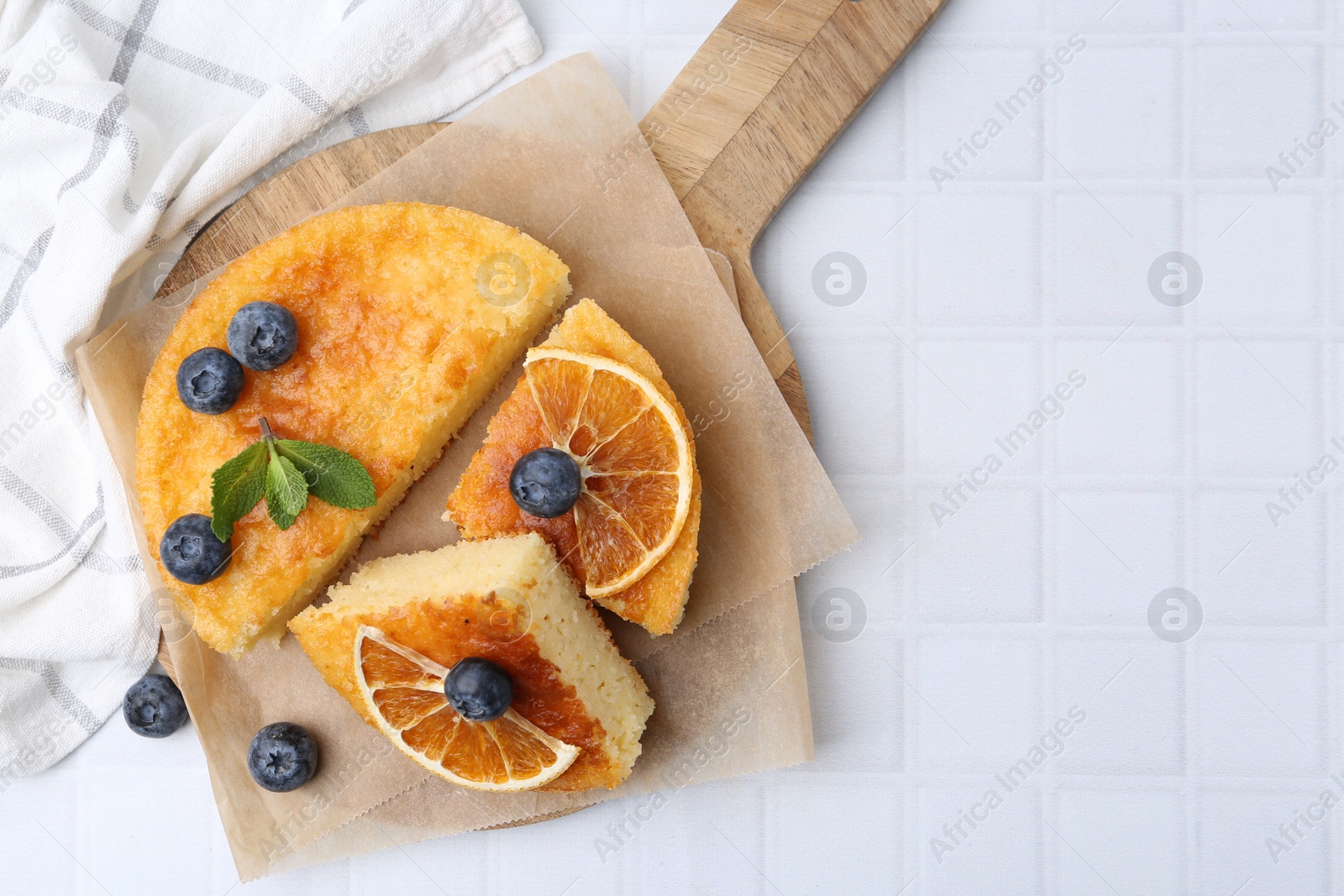 Photo of Pieces of delicious semolina cake with blueberries and orange slices on white tiled table, flat lay. Space for text