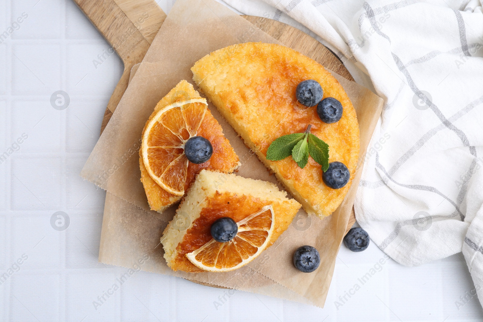 Photo of Pieces of delicious semolina cake with blueberries and orange slices on white tiled table, flat lay